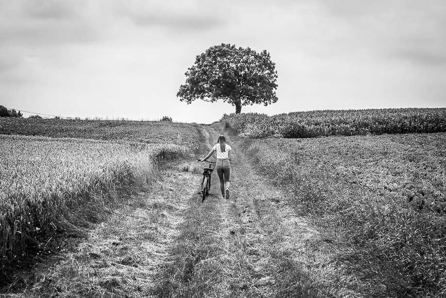 Zwart-wit beeld van een pubermeisje naast haar fiets, ze wandelt weg op een landweg in de richting van een eenzame boom.