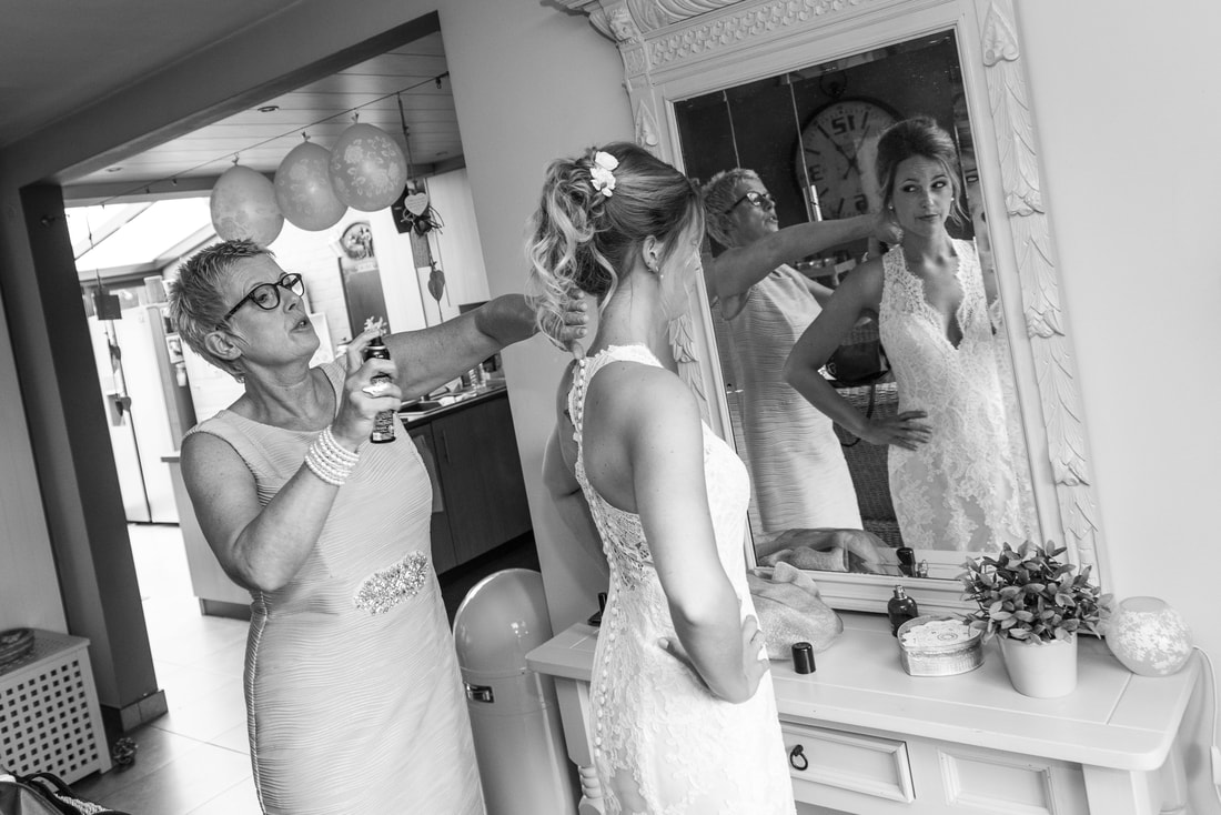 Zwart-wit foto van een bruid die zich klaarmaakt voor de spiegel, haar moeder spuit haarlak.