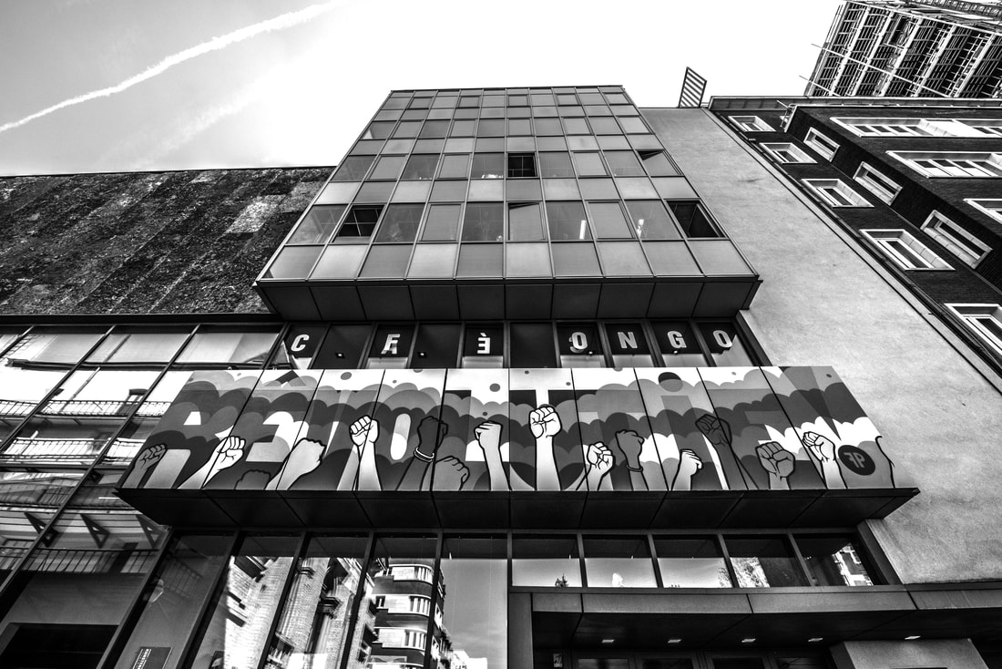 Zwart-wit foto van het KVS gebouw in Brussel, vanuit kikkerperspectief, met de focus op Café Congo en de muurtekening met vuisten.